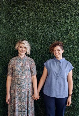 Bir gülümseyen genç lezbiyen çift ayakta el yapraklı bir duvarın dışında önünde el birlikte portresi