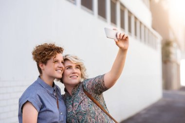 Genç lezbiyen çift ayakta kol kola selfies birlikte bir gün dışarı tadını çıkarırken alarak şehir bir sokakta gülümseyen