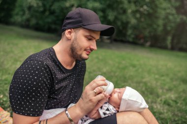 Erkek bebek onun kollarında tutarak ve onu bir formül ile dışarıda otururken bir parkta biberon bakmakta genç baba