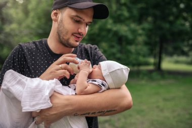 Onun bebek çocuk onun kollarında kucaklarken ve onu bir parkta dışında dururken formül şişeyle besleme genç baba bakmakta