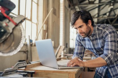 Ağaç işleme stüdyosunda bir dizüstü bilgisayar ile online çalışma odaklı woodworker bir tezgah üzerinde bir gönye ile eğilerek gördüm