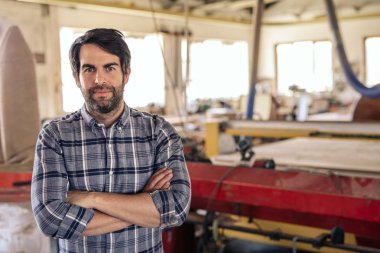 Portrait of a carpenter standing with his armscrossed in his large woodworking studio full of tools and equipment clipart