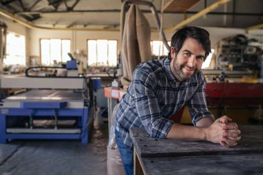 Büyük ağaç işleme stüdyosunda araçları ve ekipman dolu bir tezgah üzerine eğilerek gülümseyen bir woodworker portresi