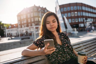 Dışında bir kahve içme ve cep telefonunu bir mesaj okuma kampüste bir bankta oturan genç Asyalı üniversite öğrencisi