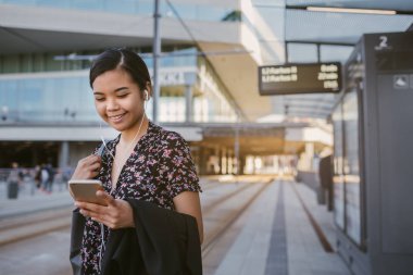 Kulaklıklar üzerinde müzik dinlemek ve onu sabah sırasında bir otobüs durağında beklerken bir metin okuma Asya öğrenci gidip genç gülümseyen