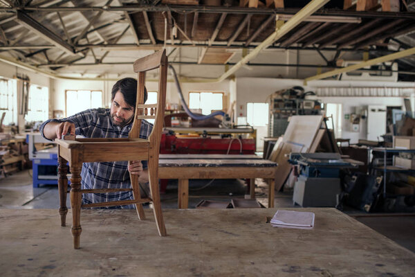 Skilled furniture maker sanding a wooden chair on a workbench while working alone in his large woodworking shop