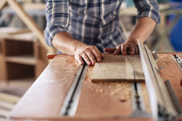 Closeup of a carpenter using a table saw to cut a plank of wood while working in his woodworking studio