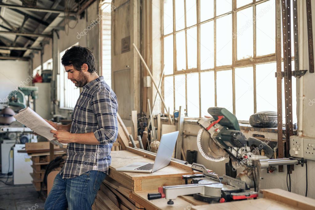 Young woodworker reading through a notebook of designs while leaning against a workbench in his carpentry studio