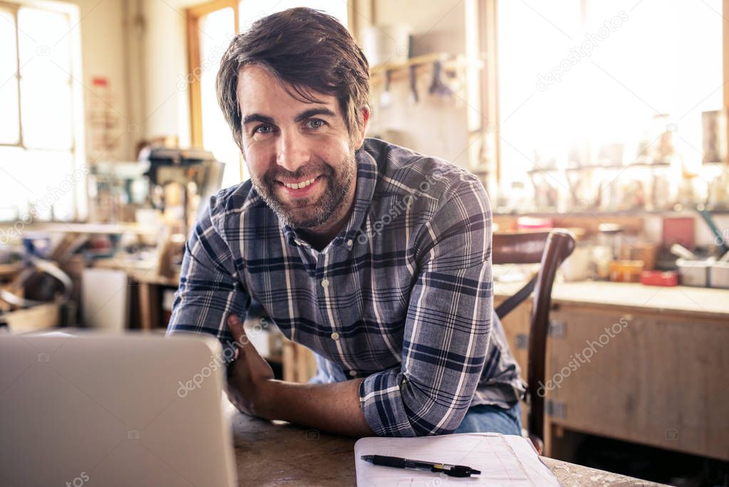Portrait of a woodworker smiling while sitting at a workbench in his carpentry studio working on a laptop