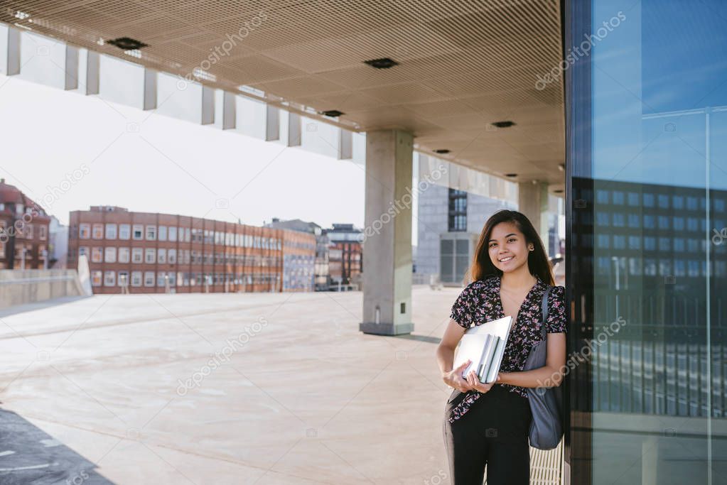 Portrait of a young Asian university student smiling confidently while leaning on a campus building holding textbooks and a laptop