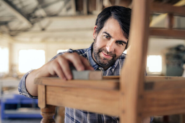 Closeup of a smiling woodworker using sandpaper to sand a wooden chair while working in his furniture making shop