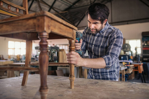 Skilled young woodworker using a drill on a wooden chair on a workbench while working alone in his large woodworking shop
