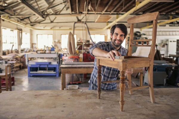 Smiling furniture maker sanding a wooden chair on a workbench while working alone in his large woodworking shop