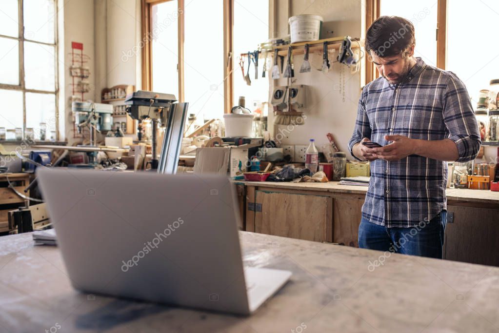 Young woodworker reading reading a text message on his cellphone and working on a laptop while standing in his woodworking studio