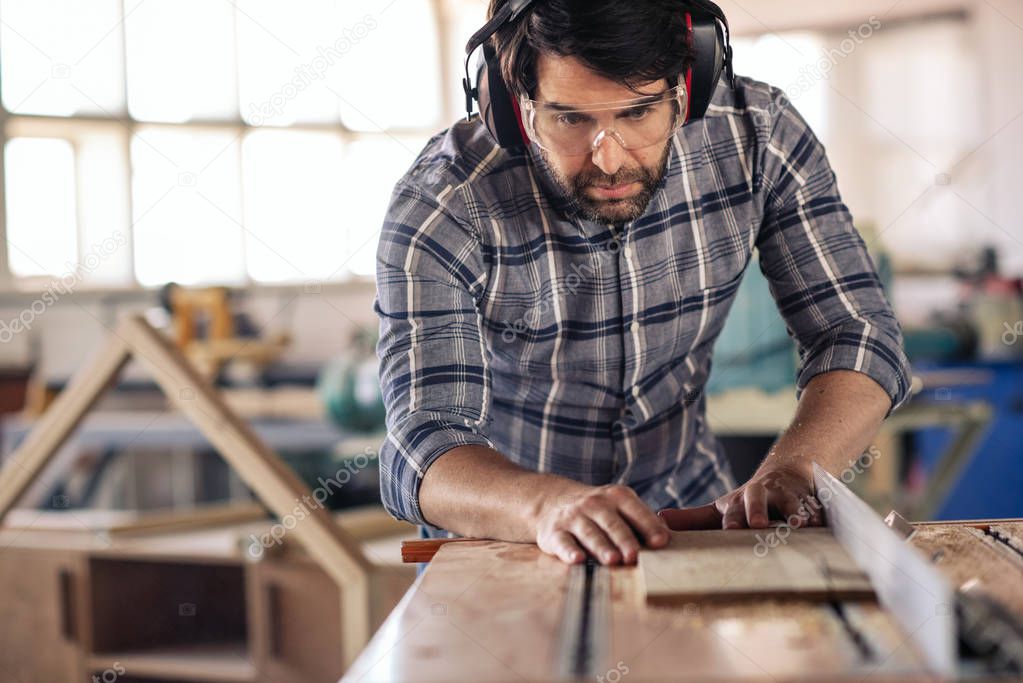 Focused carpenter wearing safety gear making precision cuts on a plank of wood using a table saw while working in his woodworking studio