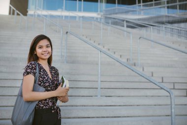 Kampüs merdivenlerde tarafından ders kitapları dururken tutarak bir gülümseyen genç Asya Kolej Öğrenci portresi
