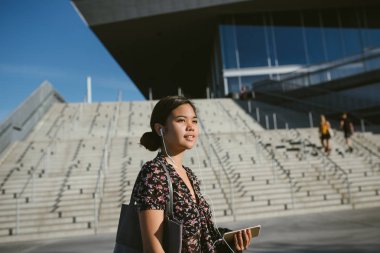Gülümseyen Genç Asyalı üniversite öğrencisi kampüste kulaklık müzik dinlerken sınıflar arasındaki yürüyüş  
