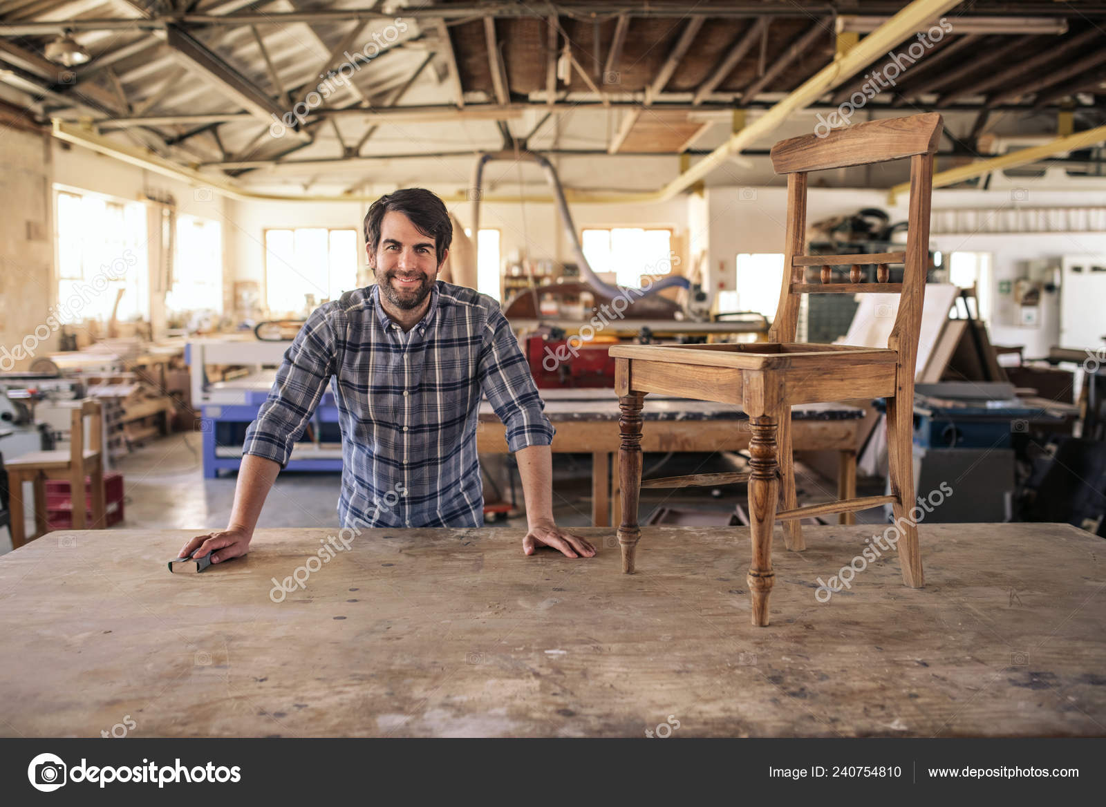 一个微笑的家具制造商的肖像靠在长凳上在他的木工工作室工作的椅子上 图库照片 C Mavoimages