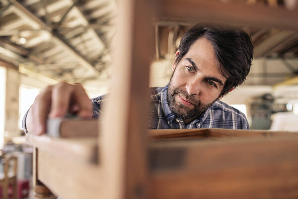 Closeup of a furniture maker using sandpaper to smooth a wooden chair while working in his carpentry shop