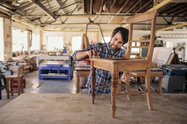 Furniture maker skillfully sanding a wooden chair on a workbench while working alone in his large woodworking shop