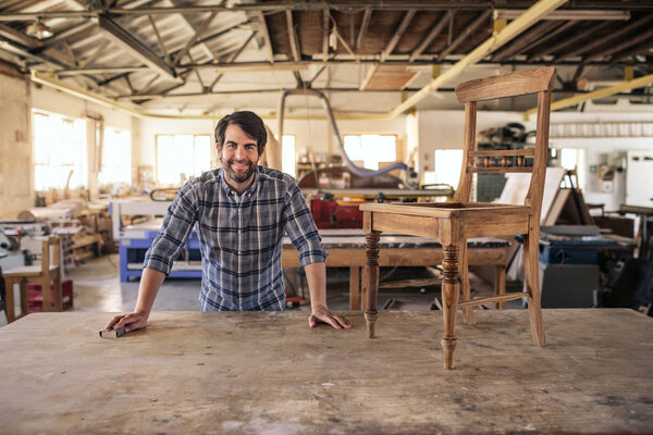 Portrait of a smiling furniture maker leaning on a bench in his woodworking studio working on a chair 