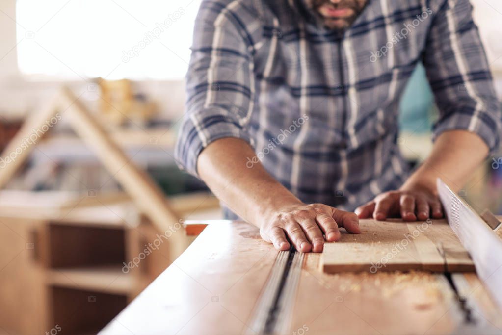 Closeup of a carpenter sawing a plank of wood with a table saw while working alone in his woodworking studio