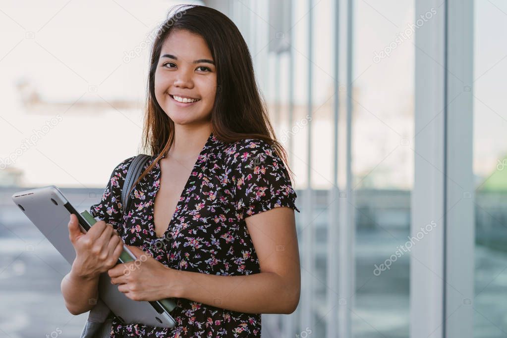 Closeup of a young Asian university student smiling confidently while standing on campus holding textbooks and a laptop