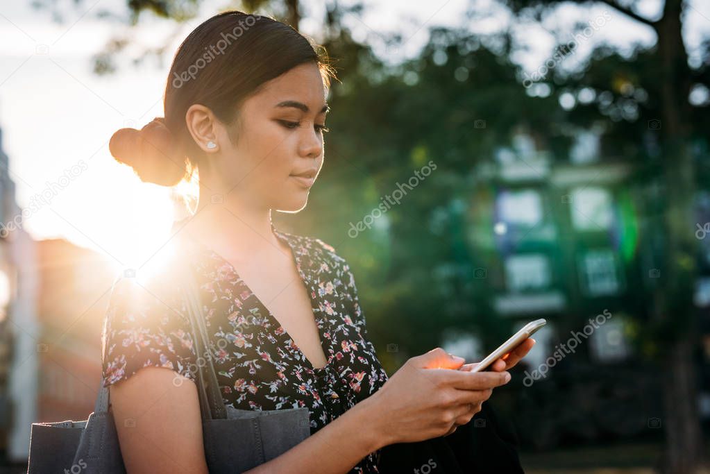 Young Asian businesswoman standing outside on a late sunny afternoon sending a text message on her cellphone