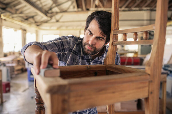 Skilled furniture maker sanding a wooden chair on a workbench while working alone in his large woodworking shop