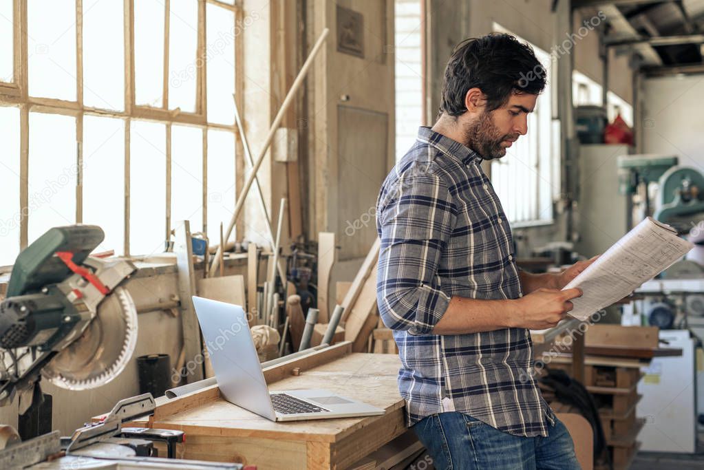 Craftsman looking through a notebook of designs while leaning against a workbench in his carpentry studio 