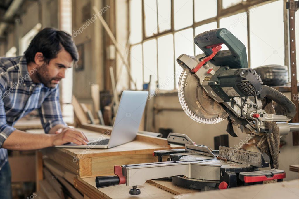 Young woodworker working online with a laptop while leaning on bench by a mitre saw in his workshop