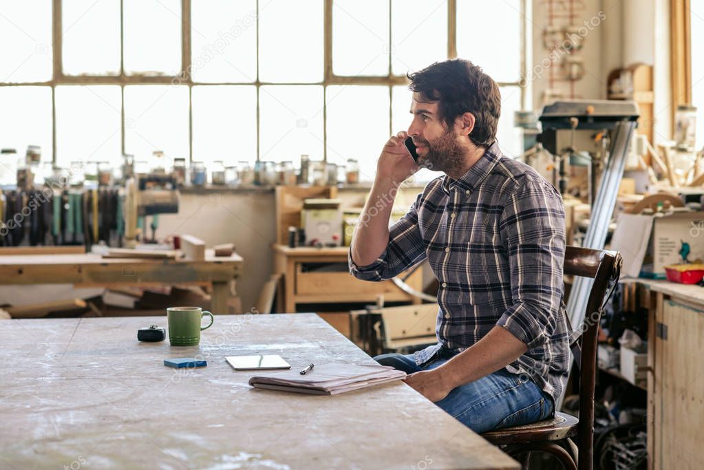 Young woodworker having a conversation on a cellphone while sitting at a bench in his woodworking studio