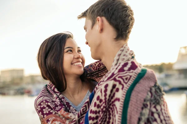 秋の午後に港で一緒に毛布に包まれながらお互いを探している若いカップルの笑顔 — ストック写真