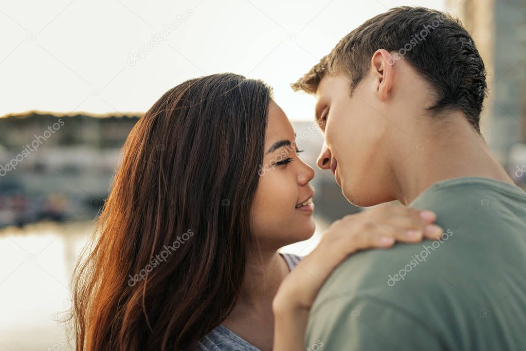 Affectionate young couple about to kiss while standing arm in arm together by a harbor in the late afternoon