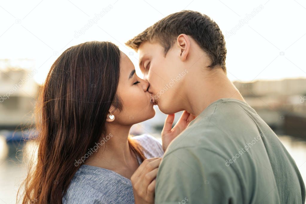 Affectionate young couple sharing a kiss while standing arm in arm together by a harbor in the late afternoon