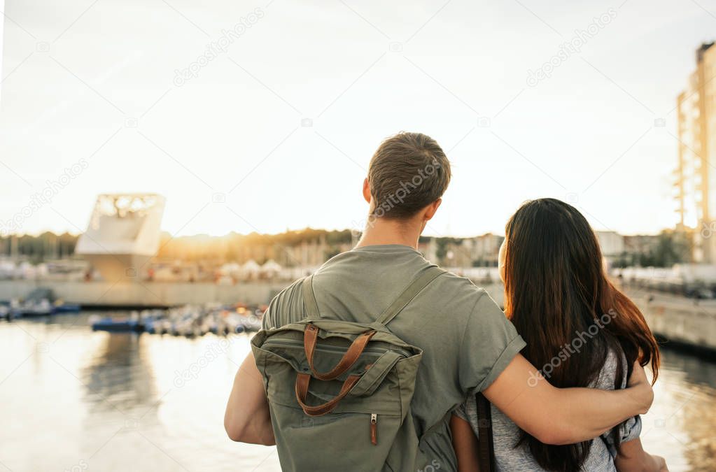 Rearview of a young couple standing arm in arm together looking out over a harbor in the late afternoon