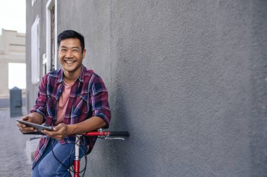 İle Onun Bisiklet dijital tablet kullanma şehirde bir duvara yaslanmış bir ekose gömlek gülümseyen genç bir Asyalı adam portresi
