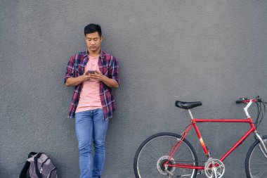 Genç Asyalı adam ekose gömlekli cep telefonunu ile Onun Bisiklet şehirde bir duvara yaslanmış iken üzerinde bir ileti gönderme