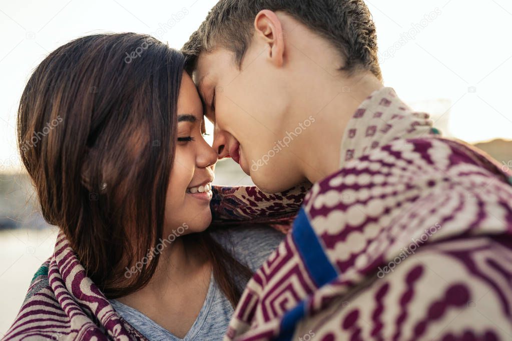 Romantic young couple about to kiss while wrapped in a blanket together by a harbor on an autumn afternoon