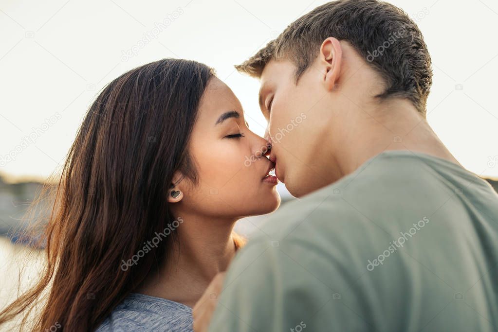 Romantic young couple kissing each other while standing arm in arm together by a harbor in the late afternoon