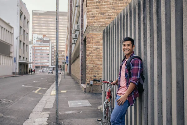 都市の壁に彼の自転車に傾いた格子縞のシャツの笑顔若いアジア人男性の肖像画 — ストック写真