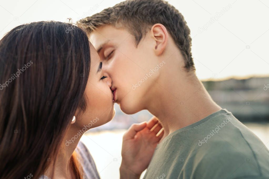 Affectionate young couple sharing a romantic kiss while standing arm in arm together by a harbor in the late afternoon