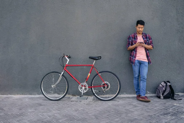 彼の携帯電話でメッセージを読んで都市の壁に彼の自転車に傾いた格子縞のシャツの若い男性 — ストック写真