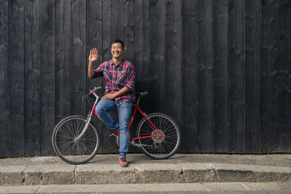 街の暗い壁の前で彼の自転車で立っている間に手を振っている笑顔の若いアジア人の肖像 — ストック写真