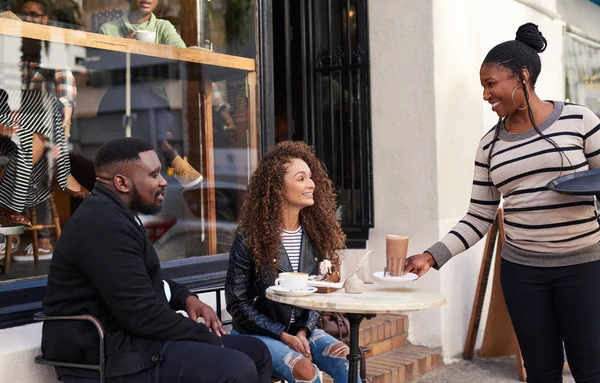 为两个面带微笑的年轻朋友在人行道上的一家咖啡馆的桌子旁准备饮料的友好女服务员 — 图库照片