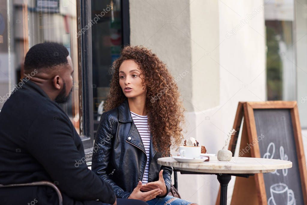 Two young friends talking together while sitting at a table outside at a sidewalk cafe having drinks and a snack