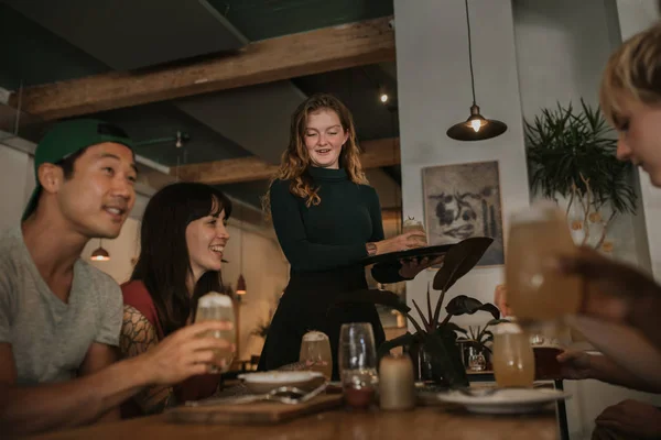 Smiling waitress bringing a drinks order to a group of young friends enjoying a night out together in a bar