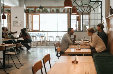 Popüler bir bistroda bir masada öğle yemeği ve içecekler üzerinde konuşurken gülümseyen çeşitli arkadaşlar İçerik grubu