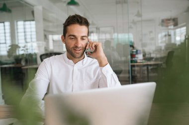 Gülümseyen genç işadamı bir dizüstü bilgisayar üzerinde çalışan ve modern bir ofiste masasında otururken bir cep telefonu konuşuyor