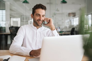 Modern bir ofiste masasında bir dizüstü bilgisayar üzerinde çalışırken onun cep telefonu konuşurken Gülümseyen genç işadamı